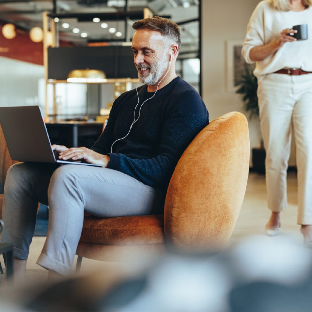 A man with headphones is sitting comfortably in a modern coworking space—One Two Five by Hub— smiling as he works on his laptop. He is seated on an orange armchair, dressed in casual attire, while a woman in the background walks by holding a coffee cup. The space features stylish furniture, warm lighting, and an open, contemporary design.