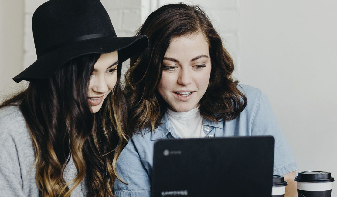 two young women huddled around laptop, looking at screen.
