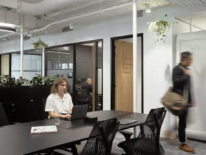 a woman at hot desk typing on a computer and a man walks past