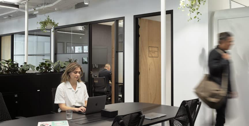 a woman at hot desk typing on a computer and a man walks past