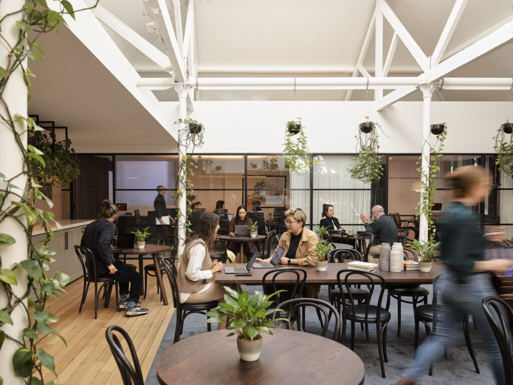 A café setting found in an office with people sitting at tables. There are plants hanging from the roof.
