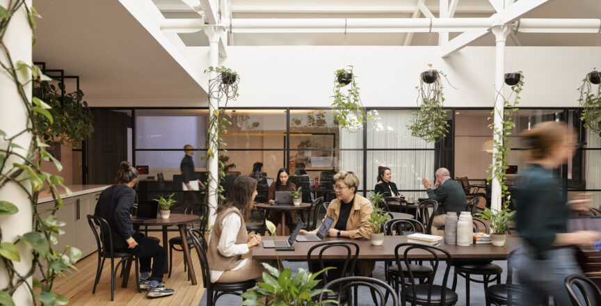 A café setting found in an office with people sitting at tables. There are plants hanging from the roof.