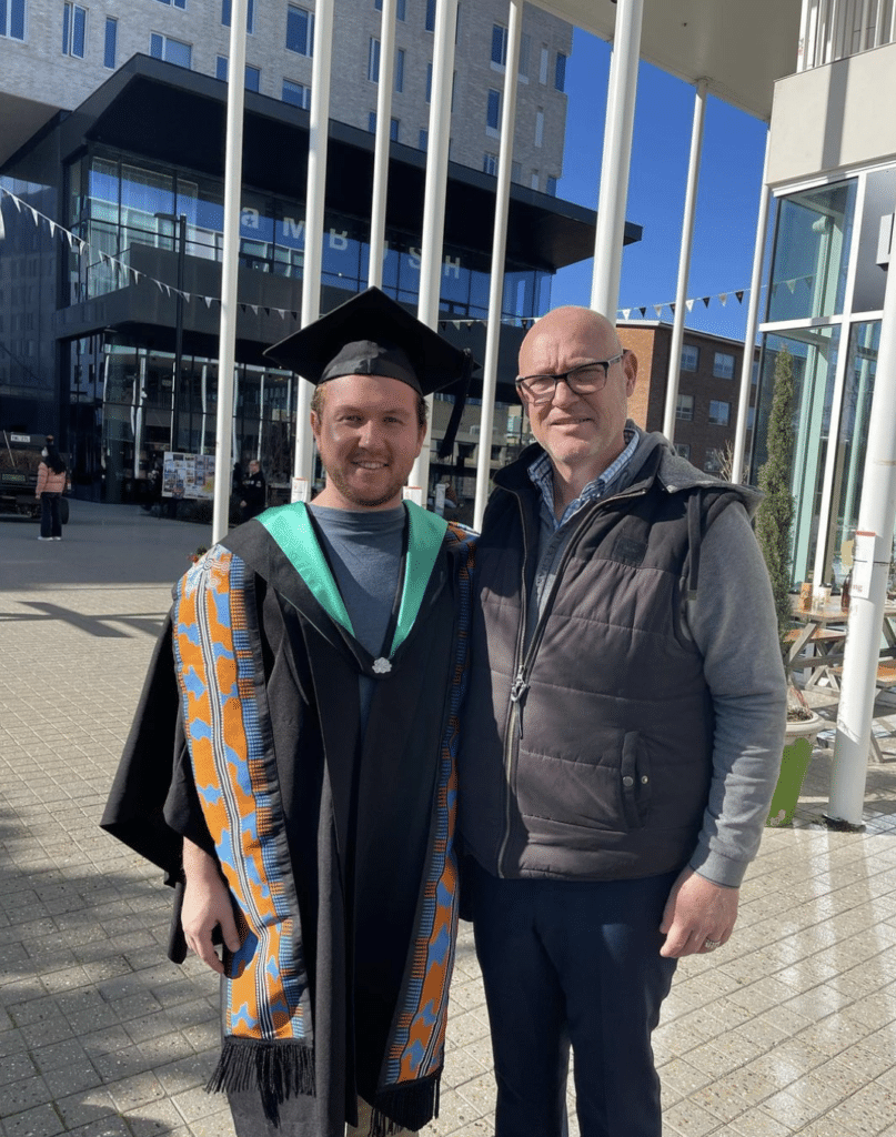 two men standing next to eachother, one has graduation clothes on