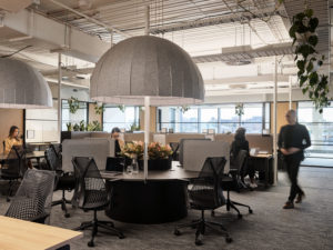 A shared office space featuring a range of tables. There is a woman sitting at a desk on her laptop.
