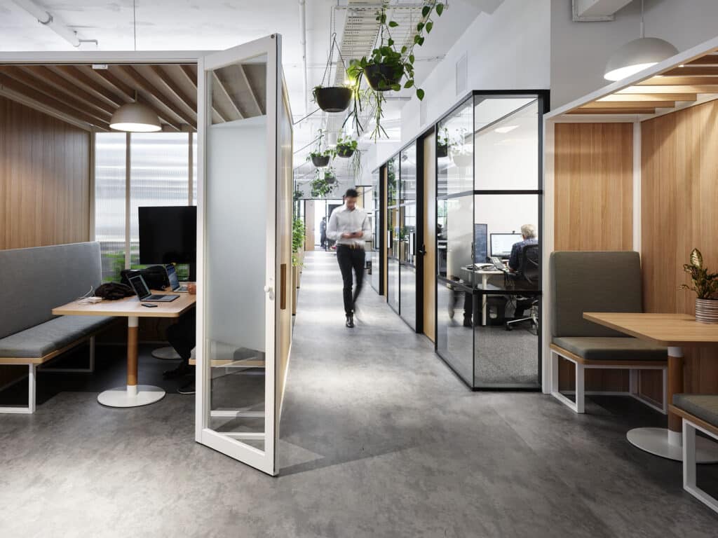 a man walks down a hallway in an office. there are plants hanging over head and a range of benches and seating options around.