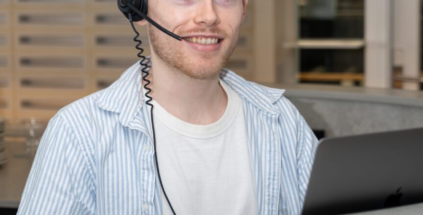 Man sitting at a desk in front of a computer with a headset on. He's looking at the camera smiling