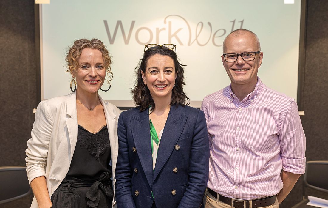 Three professionally dressed people standing and smiling in front of a 'WorkWell' signage