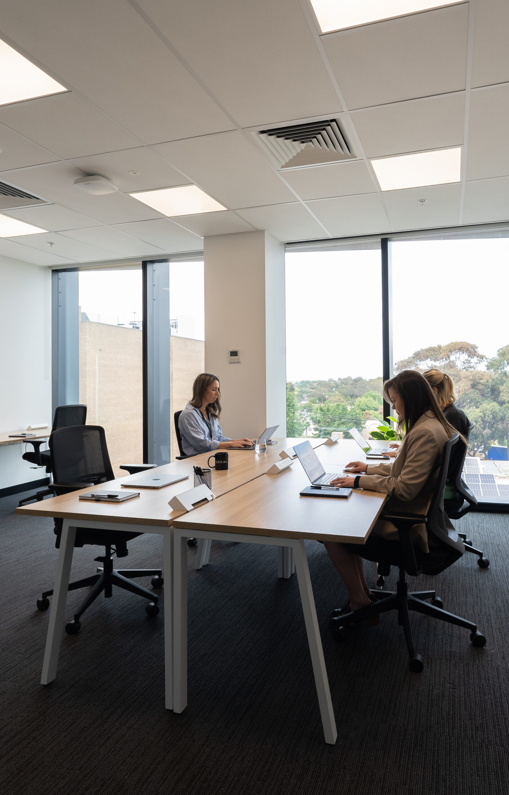 Modern office space with three people working at a large shared desk, each using laptops. The room features floor-to-ceiling windows with a view of greenery and nearby buildings. The workspace is bright, minimalistic, and equipped with ergonomic chairs.