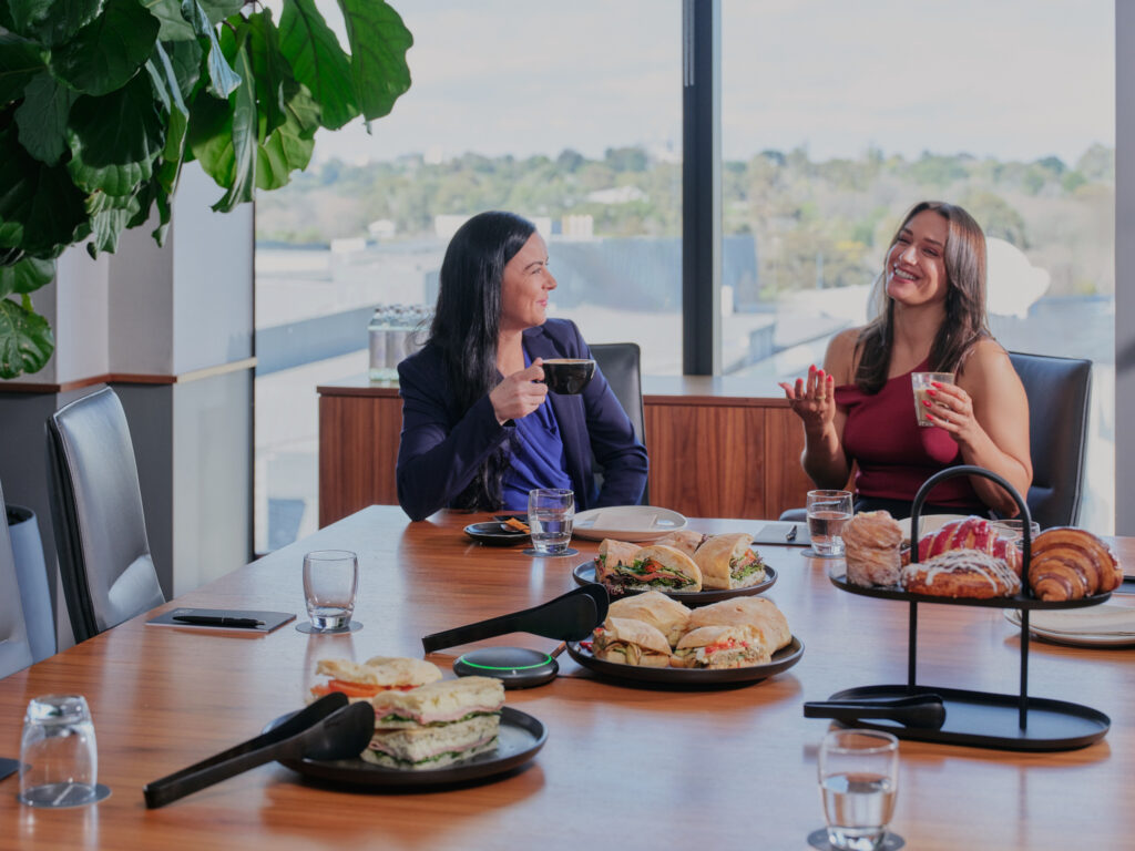 Two women in a modern boardroom at Hub Flinders Street enjoying coffee and a spread of sandwiches, pastries, and drinks, with large windows showcasing a scenic outdoor view.