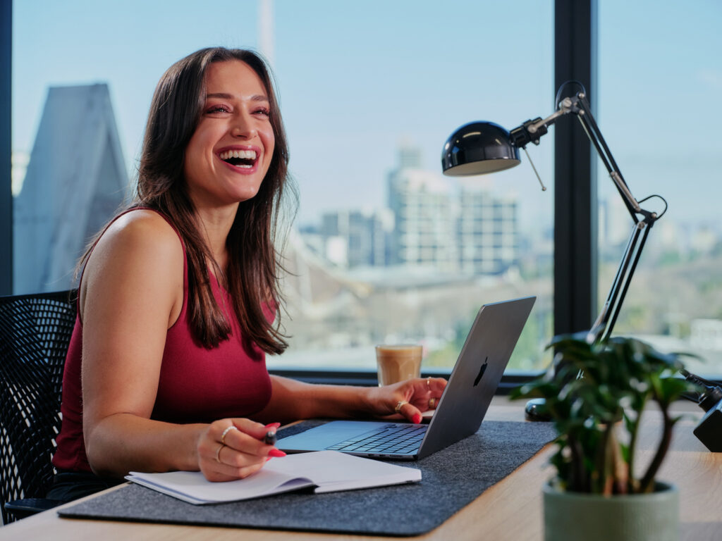 Smiling woman in a red sleeveless top working at a desk with a laptop, notebook, and coffee cup. A large window behind her shows Melbourne's cityscape view, and a desk lamp and plant are on the table.