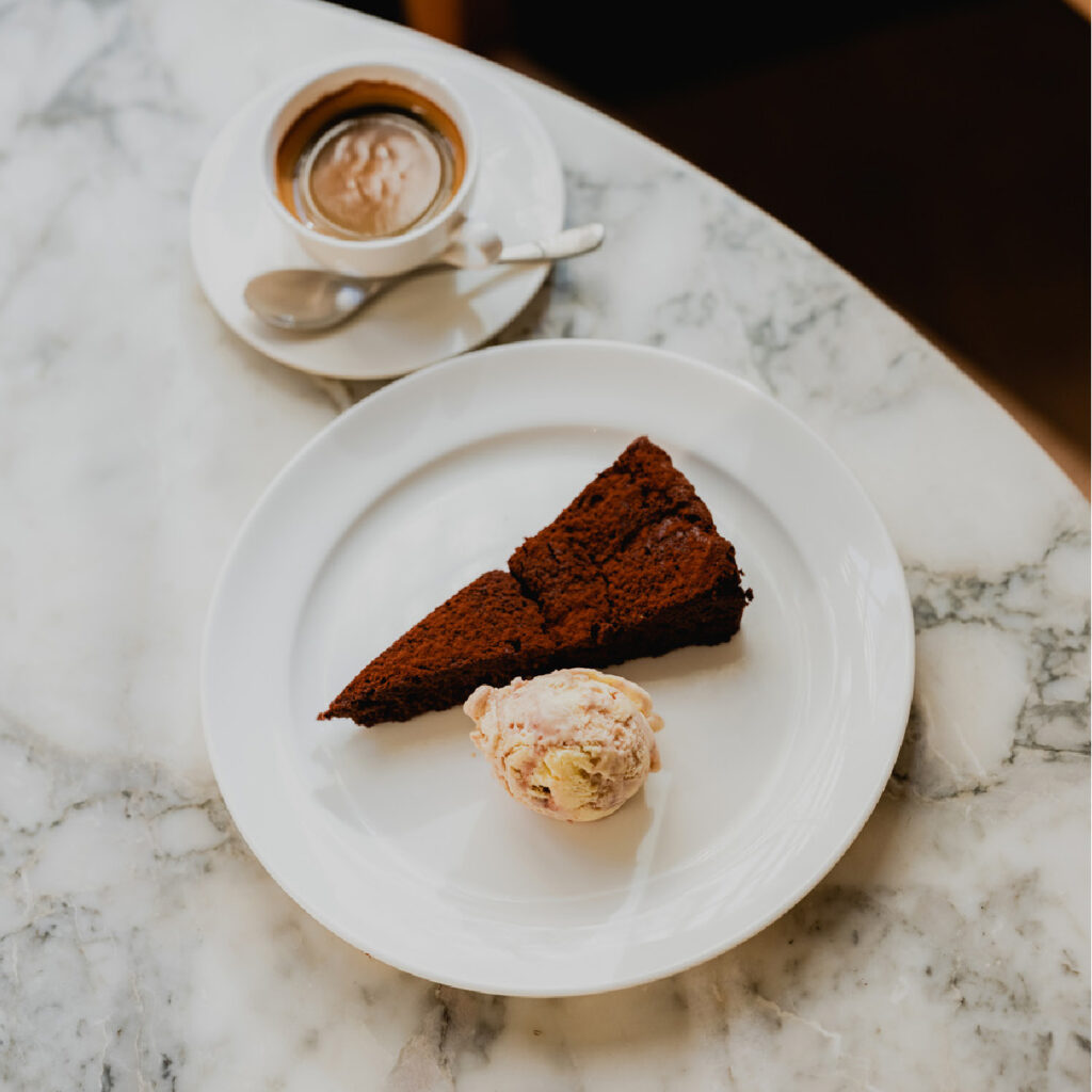 A slice of rich chocolate cake and a scoop of ice cream are beautifully plated on a white dish, sitting on a marble table. Nearby, a cup of espresso with a spoon rests on a saucer, completing the elegant One Two Five by Hub Café