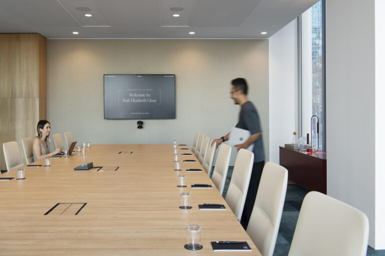 A modern boardroom with a large wooden conference table surrounded by beige swivel chairs, set against floor-to-ceiling windows with a scenic city and river view. Natural light fills the space, highlighting a sleek and professional meeting environment.