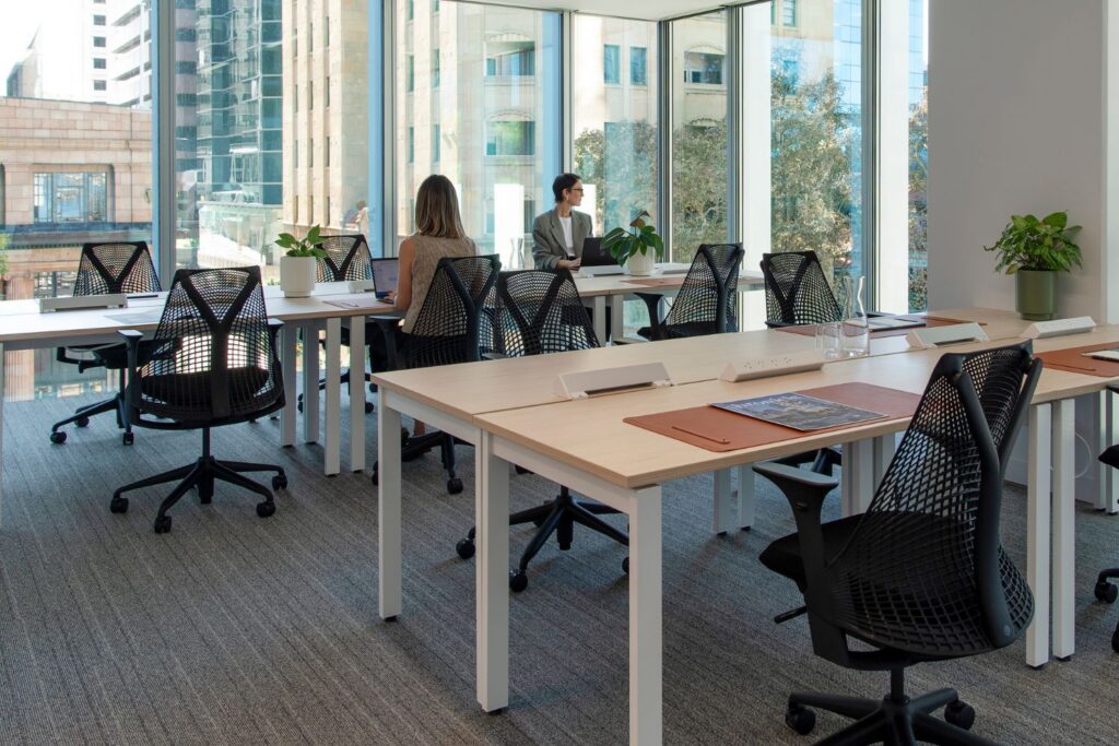 A bright, corner day office with floor-to-ceiling windows overlooking a city, featuring multiple white desks and ergonomic black mesh chairs.